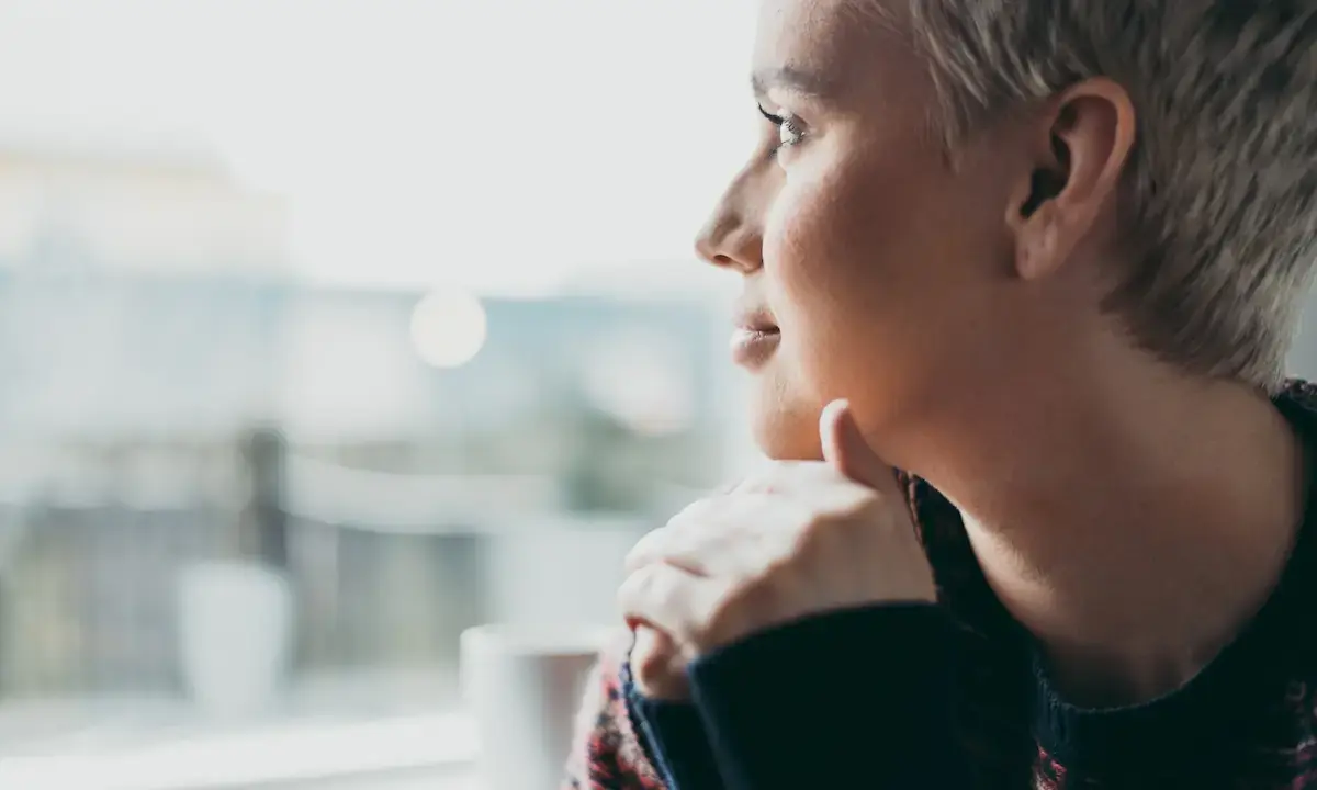 Contemplative person with short hair looking out a window, hand on chin