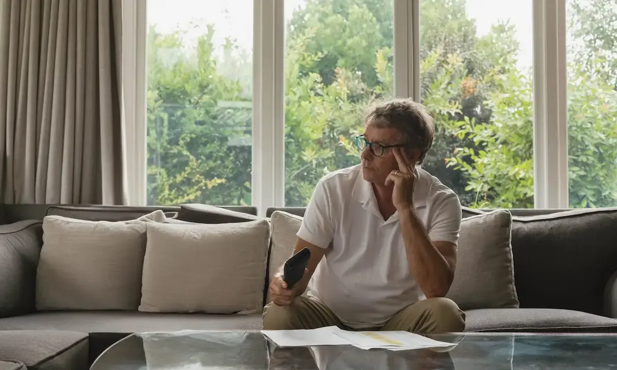 man on living room sofa holding a remote, looking thoughtful toward window